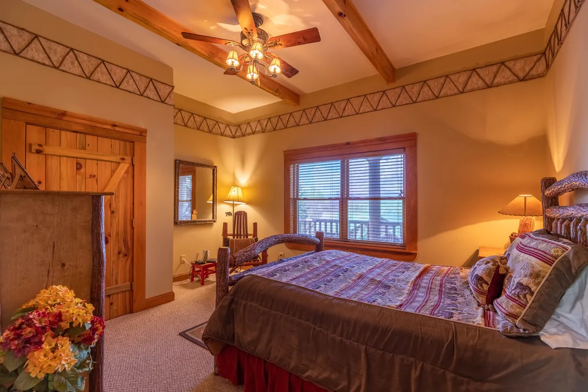 A warmly lit bedroom featuring a king bed with decorative bedding, wooden beams on the ceiling, a large window letting in natural light, a decorative mirror, and a lamp on the bedside table. The space feels restful and inviting, perfect for private relaxation.