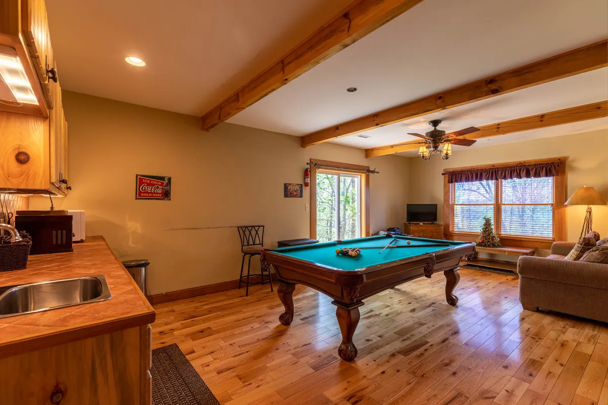 This interior shot presents a spacious lower-level entertainment area with a pool table at the center, a comfortable sofa for lounging, large windows bringing in natural light, and a kitchenette in the background. The layout encourages both active play and relaxed movie watching, capturing the essence of the family game lounge.