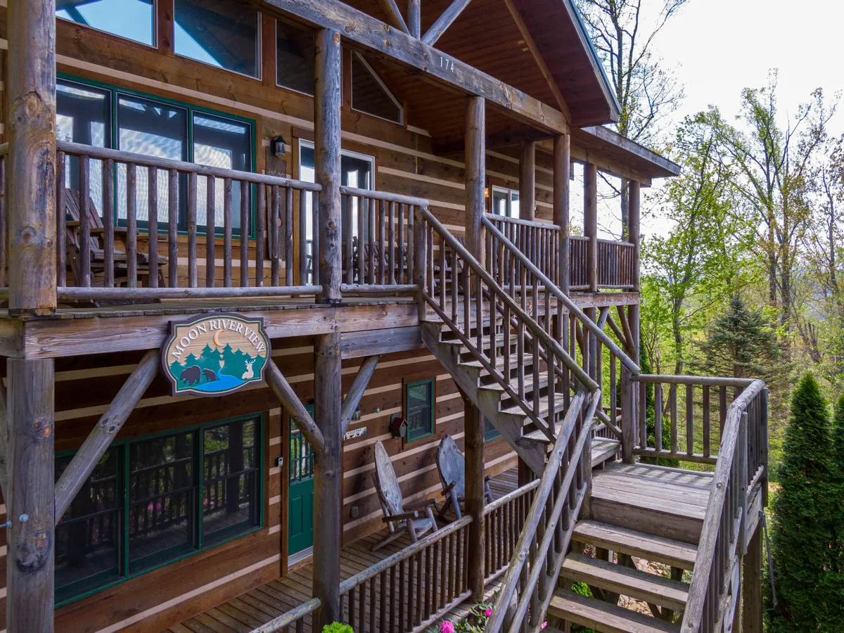 This image features the exterior of the cabin, showcasing its wooden log structure, green doors, the 'Moon River Inn' sign, and inviting rocking chairs on the porch. The cabin is surrounded by lush greenery, creating a cozy and welcoming first impression.