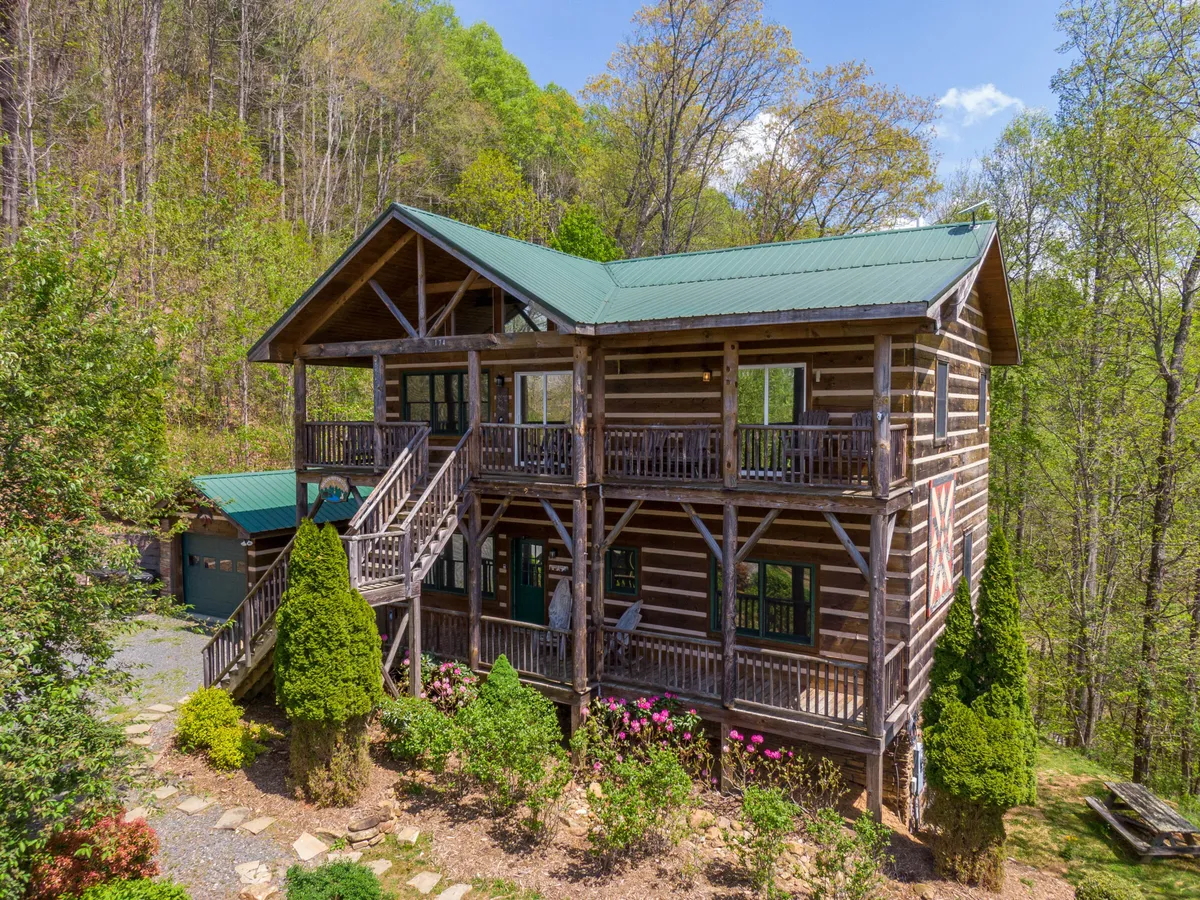 A wide exterior view of the log cabin retreat surrounded by trees and flowering shrubs, with a stone pathway leading up to the welcoming porch. The spacious grounds suggest plenty of room for outdoor exploration and relaxation.