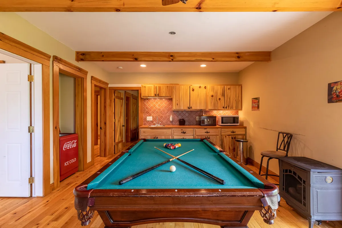 This image features a regulation-sized pool table set in a cozy game lounge, accented by wooden cabinetry and a convenient kitchenette. A stool is nearby for casual seating, and the overall ambiance is relaxed and inviting, perfect for friendly competition and family gatherings.