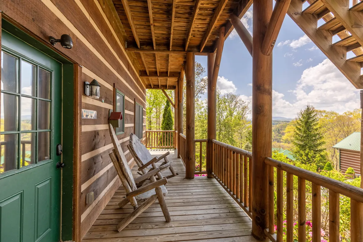 The image showcases the inviting exterior of a cabin nestled in Boone, North Carolina. Prominent features include classic wooden rocking chairs on a spacious porch, a vibrant green front door, rustic wooden railings, and a backdrop of lush natural landscape. The setting evokes a sense of tranquility, perfect for relaxing outdoors, sipping morning coffee, and soaking in the peaceful surroundings.