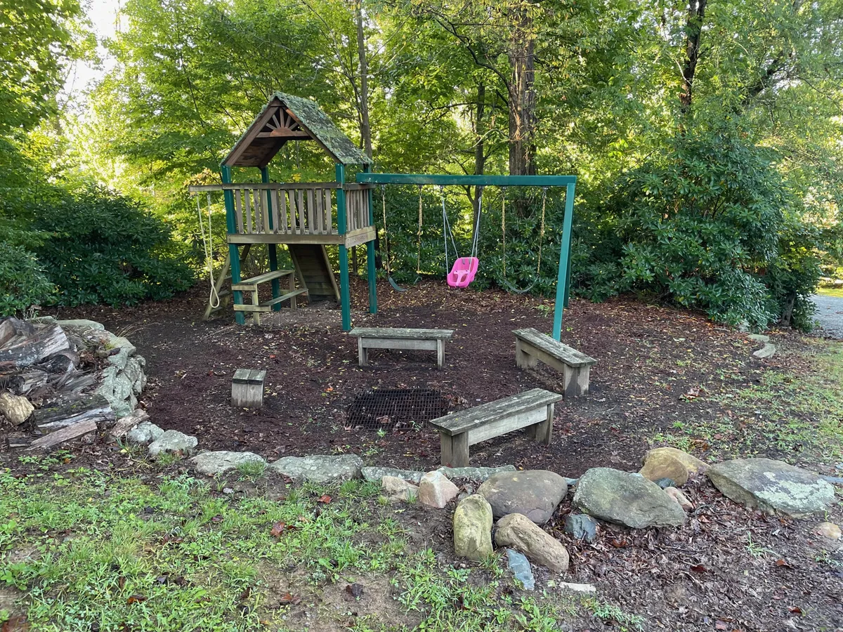 This image captures an outdoor family-friendly area featuring a playset with swings, a fire pit surrounded by benches, and open green space. The joyful, inviting mood is perfect for families looking to relax, play, and gather outdoors.