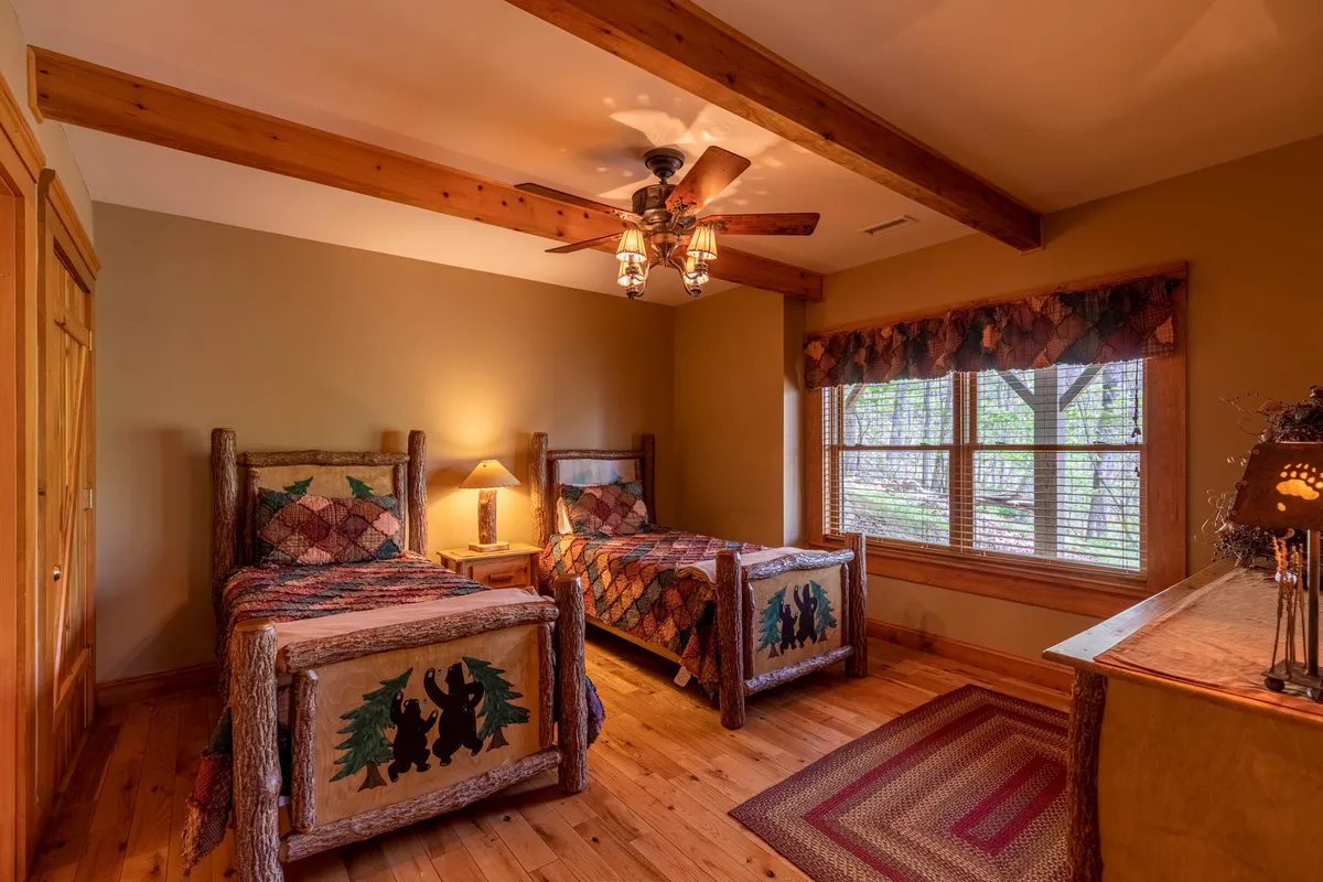 A cozy bedroom setup with twin log beds topped with quilts, a chest of drawers, a window offering nature views, and a ceiling fan. The room exudes warmth and is ideal for children or additional guests seeking a secluded sleep space.