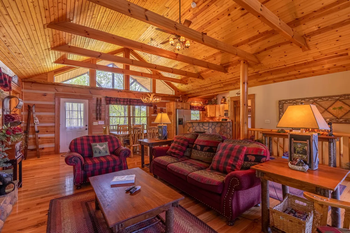 A spacious great room featuring plaid sofas arranged around a large stone fireplace, with wooden beams overhead and expansive windows letting in natural light. Decorative lamps and accents create a cozy, inviting atmosphere ideal for gatherings.