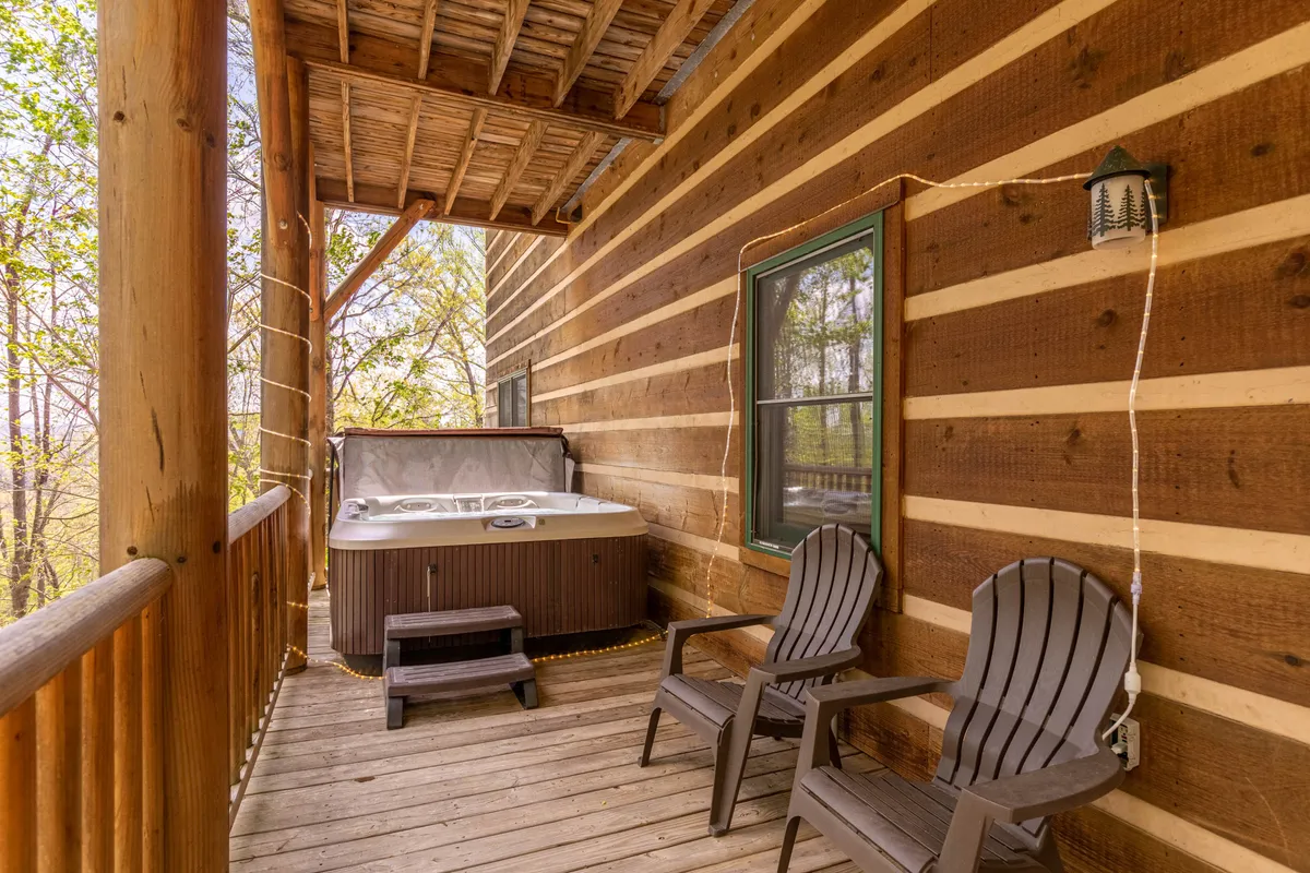 This exterior shot shows a wooden deck with a hot tub, rustic chairs, and the cabin’s log wall as a backdrop. The setting is tranquil, surrounded by trees, suggesting a perfect space for music, relaxation, and connecting with nature.