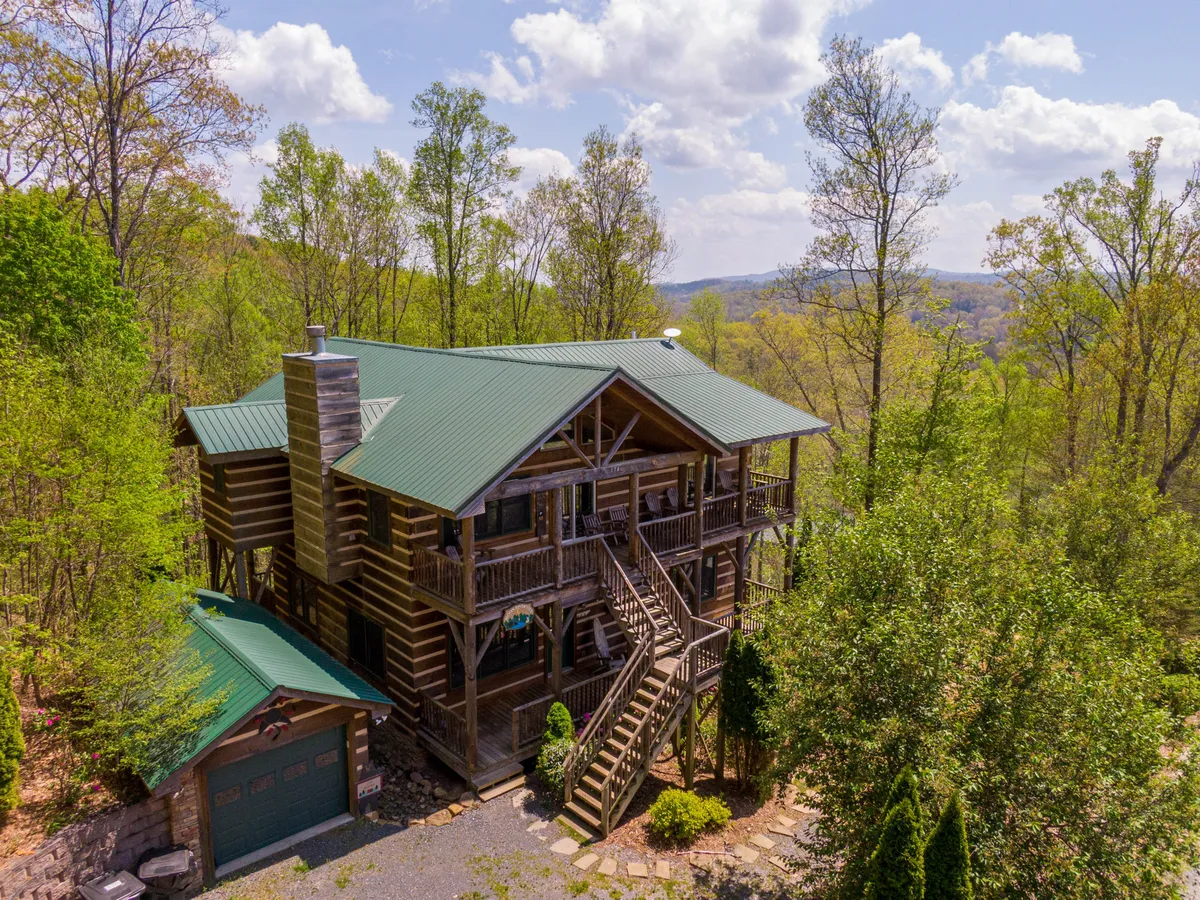 The exterior of a welcoming log cabin is pictured, with a green metal roof, lush trees, and stairs leading to a cozy porch—perfect for gatherings and alfresco dining.