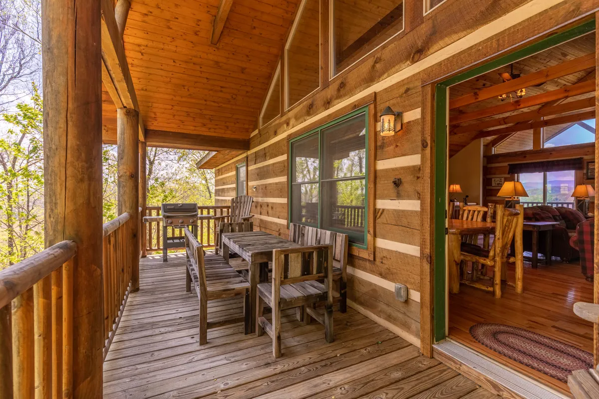 An outdoor deck with a wooden dining table and log railing, set against a backdrop of trees. The deck offers an inviting space for al fresco dining or evening relaxation, with easy access to the home's interior.