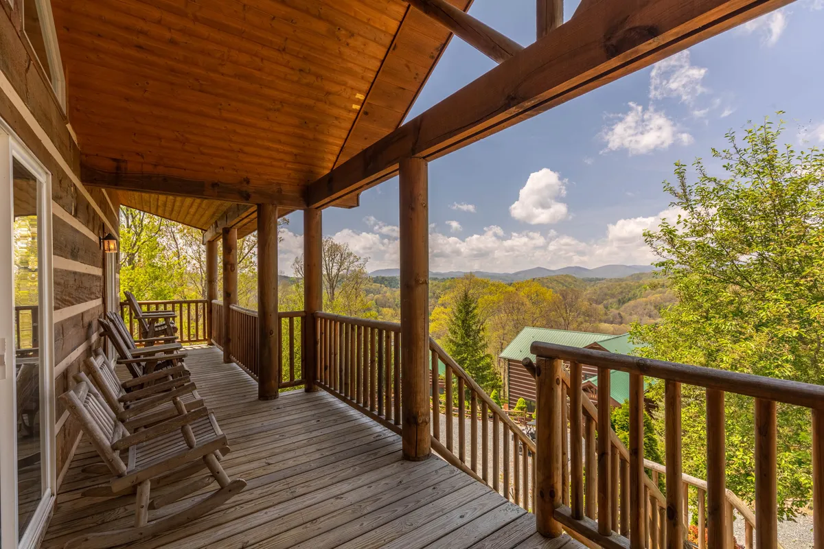 A wide wooden deck features classic rocking chairs and a railing overlooking distant mountains and lush greenery, evoking a serene and inviting ambiance.
