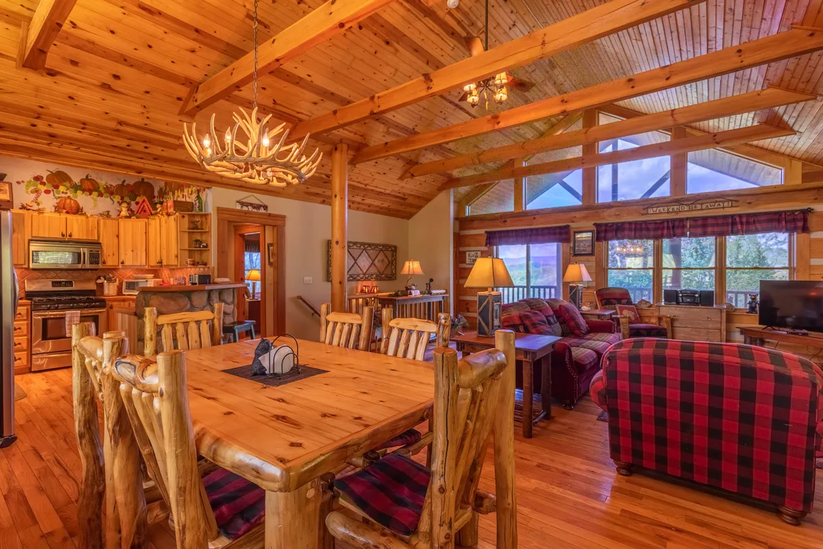 A rustic dining area with a large wooden table, antler chandelier, and comfortable seating. Large windows flood the space with natural light, and a cozy seating area by the fireplace is visible nearby, enhancing the room's inviting feel.